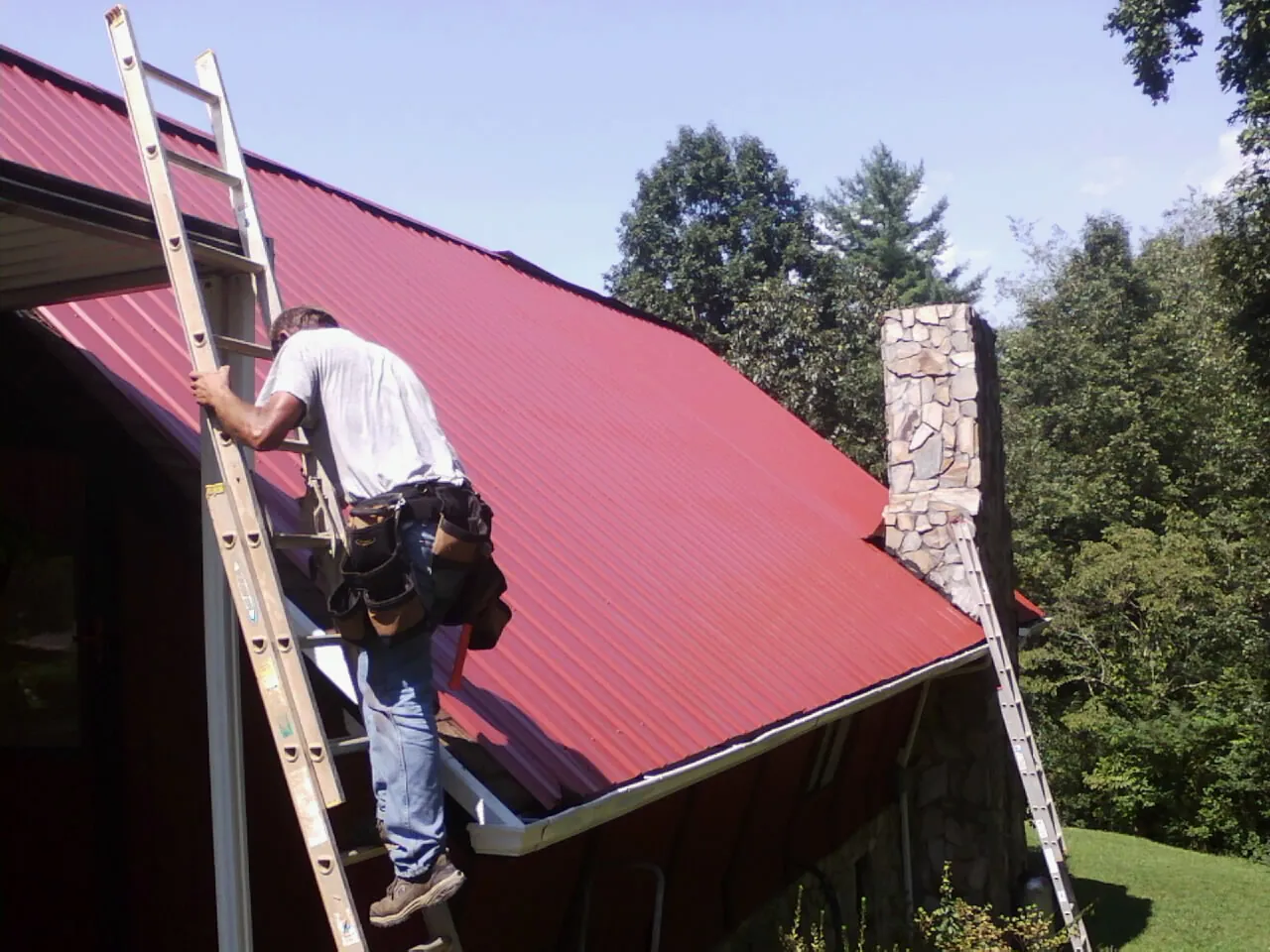 Roofer on a ladder beside a red metal roof and stone chimney — Soffit & Fascia Repair by Roofing Brothers in Mount Royal